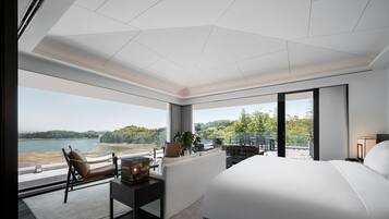 Habitación de hotel moderna con una cama grande, un área de estar con una silla y una mesa, vistas a un lago y un balcón con mobiliario exterior.