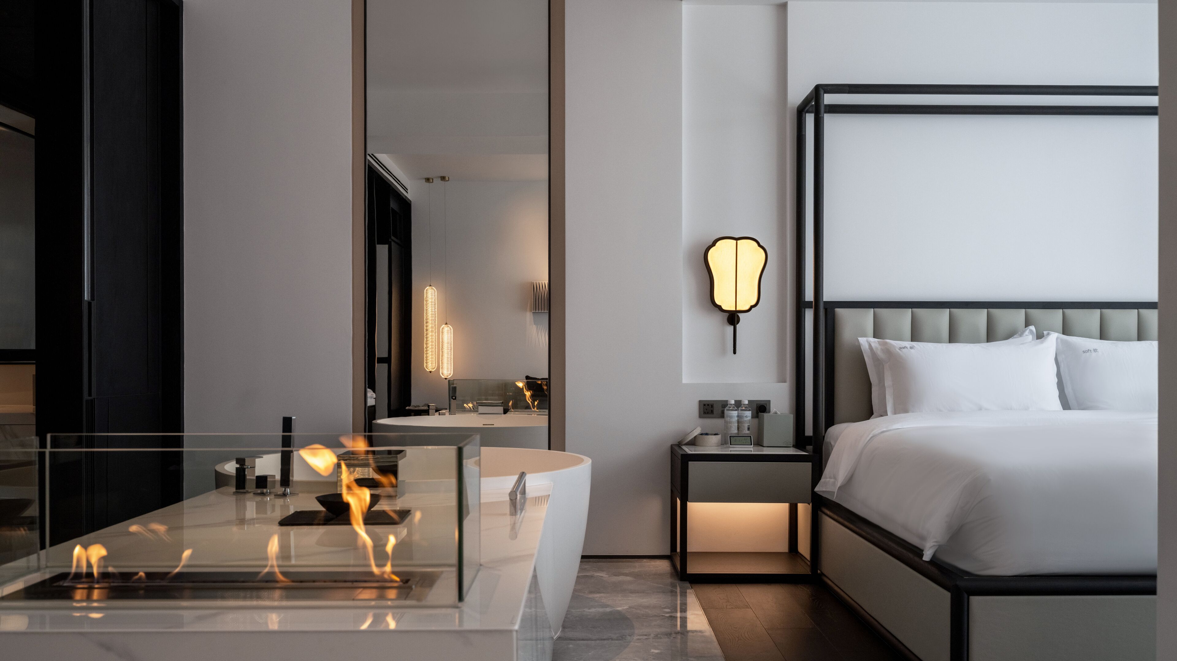 A modern hotel room with a large bed, a glass-enclosed bathtub, a bedside table with a lamp, and a mirror.