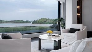 A modern living room with a large window overlooking a lake and greenery. There is a white sofa, a round coffee table with a vase of yellow flowers, and a bookshelf. - FU VALLEY (Changzhou)