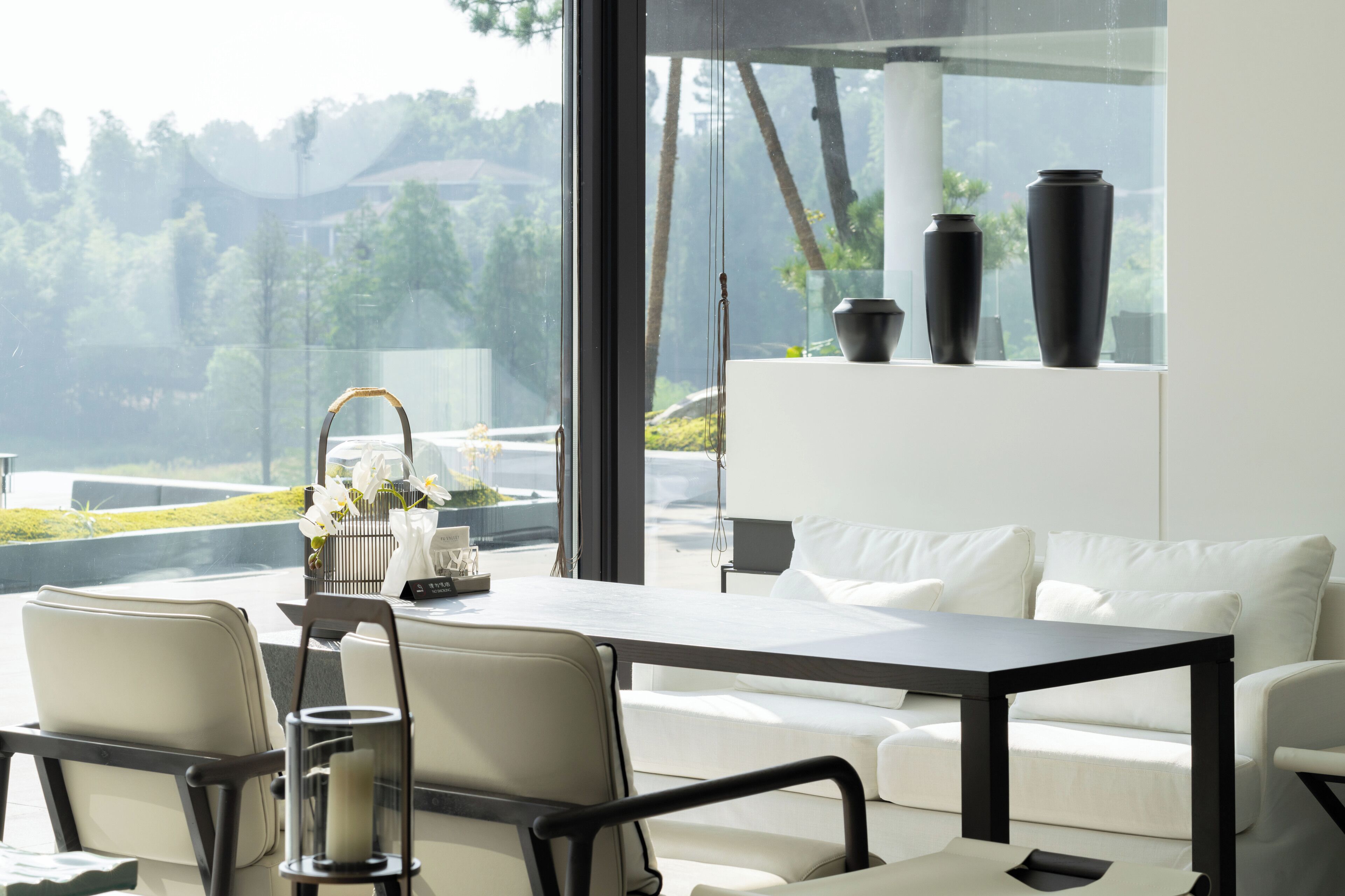 Una habitación moderna con un ventanal grande, un sofá blanco, una mesa de comedor y jarrones negros en una estantería.