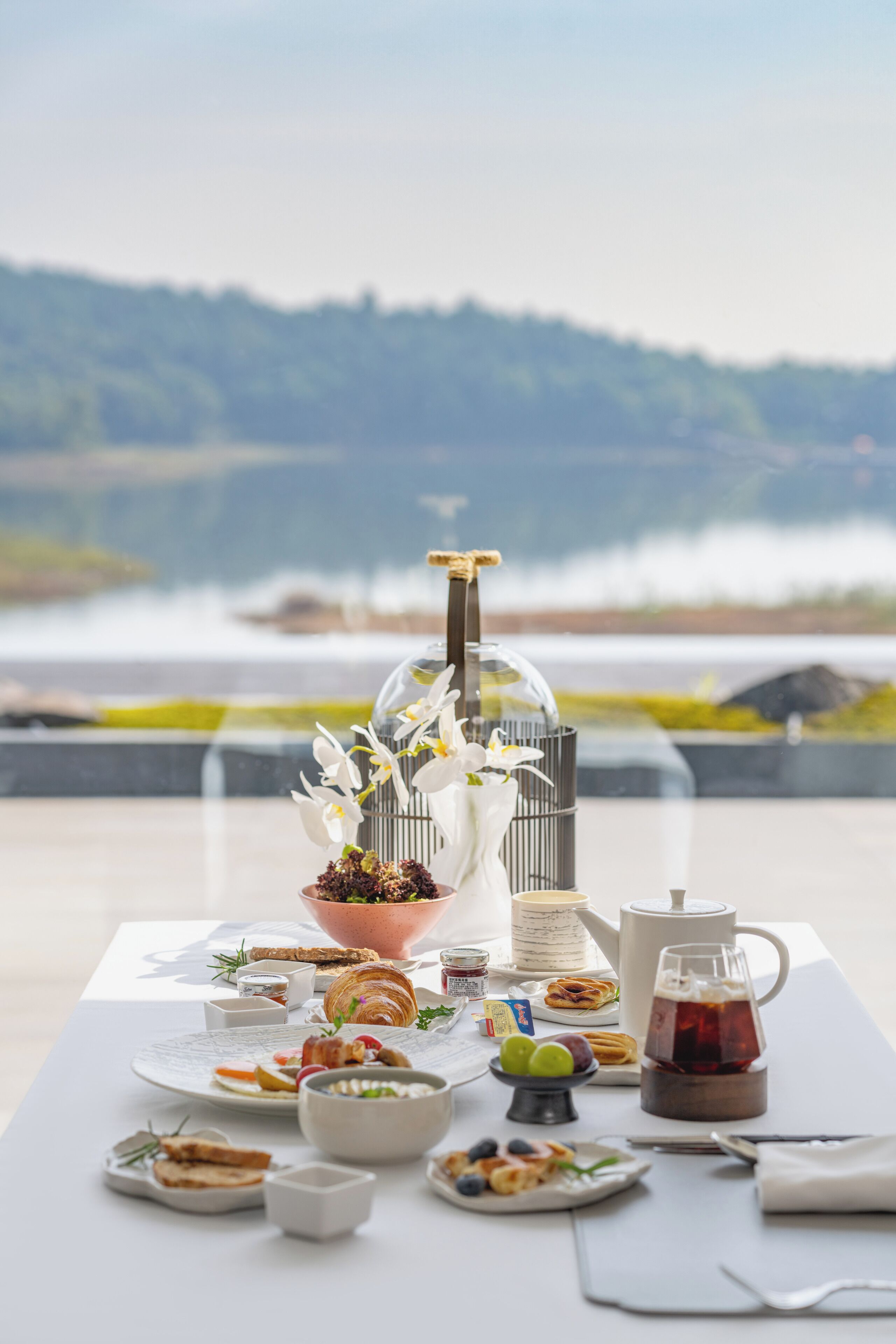 A table set with breakfast items including croissants, fruit, and tea, overlooking a lake.
