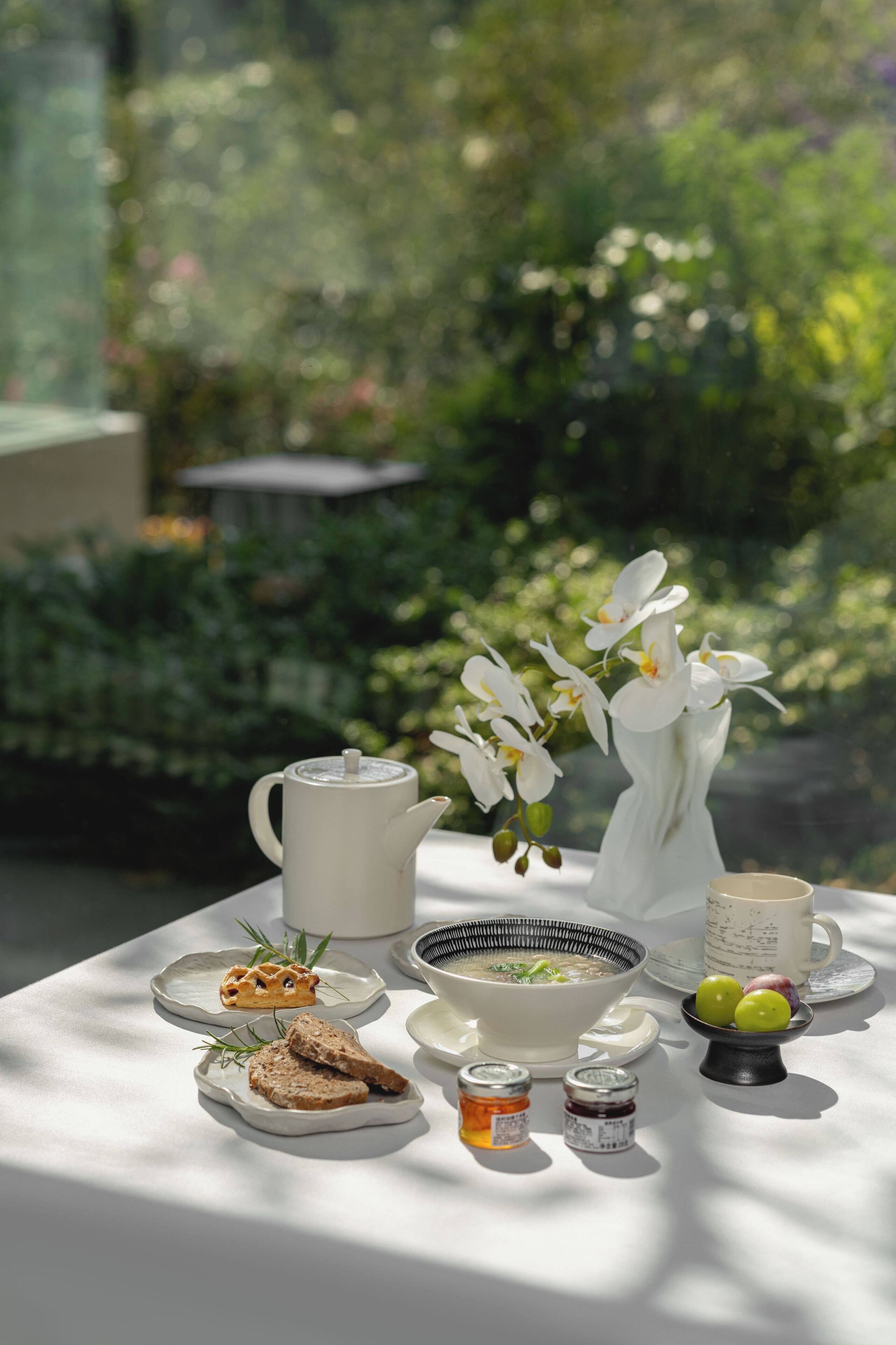 Ein gedeckter Tisch mit Teekanne, einer Suppenschüssel, Brot, Beilagen und einer Vase mit weißen Blumen.