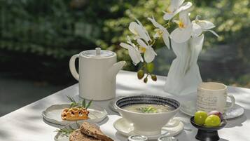 Ein gedeckter Tisch mit Teekanne, einer Suppenschüssel, Brot, Beilagen und einer Vase mit weißen Blumen.