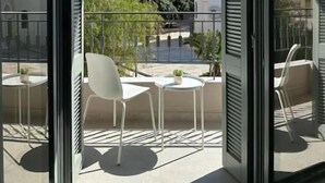 A balcony with two white chairs and a small table, overlooking a residential area with buildings and greenery. - Main Suite (Rethymno)