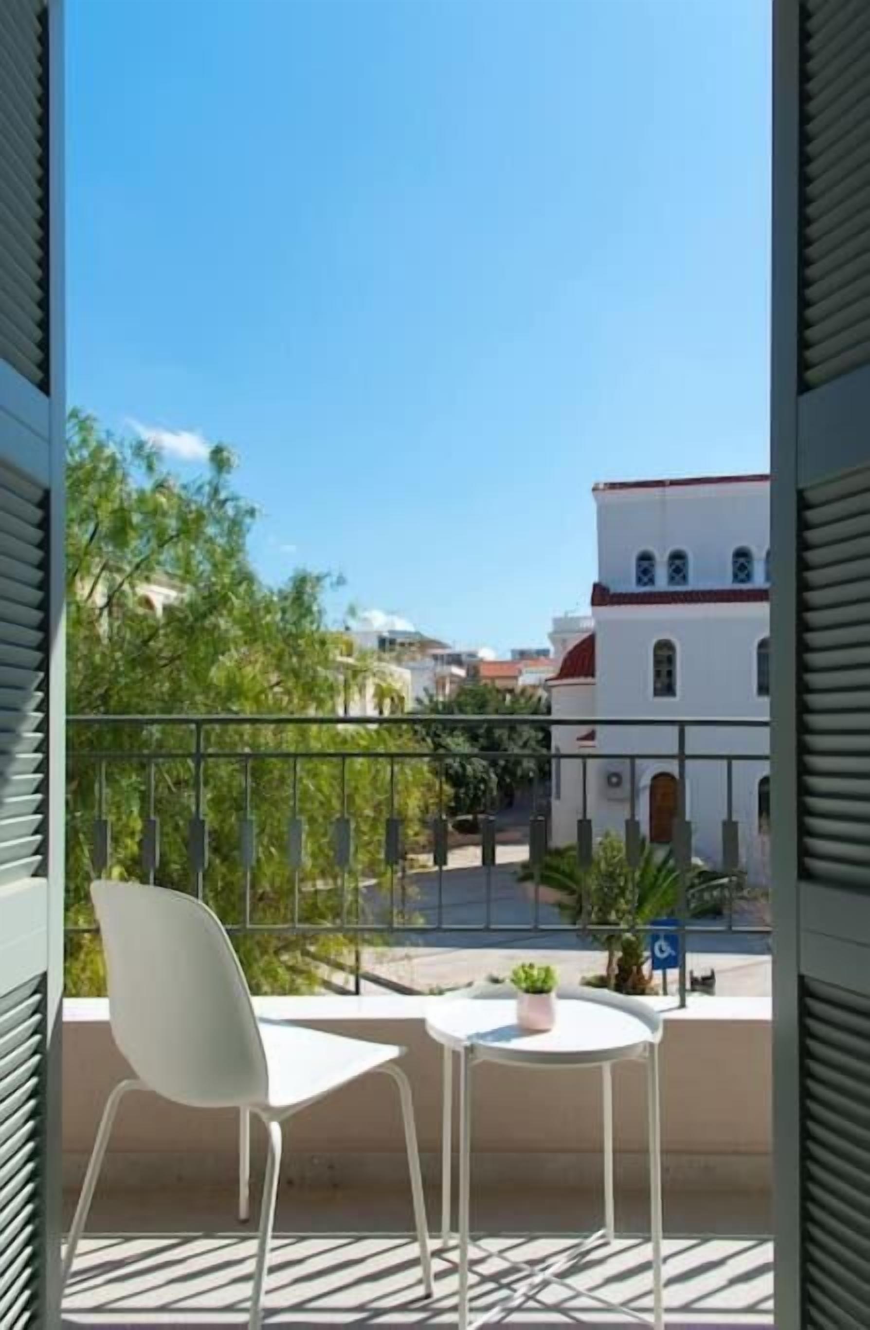 Ein Balkon mit zwei weißen Stühlen und einem kleinen Tisch, Blick auf eine Straße mit Gebäuden und blauem Himmel.