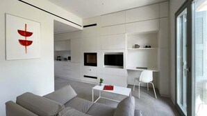 A modern living room with a grey sofa, a white coffee table, and a TV mounted on the wall. There is a dining area in the background with a kitchen visible. - Main Suite (Rethymno)