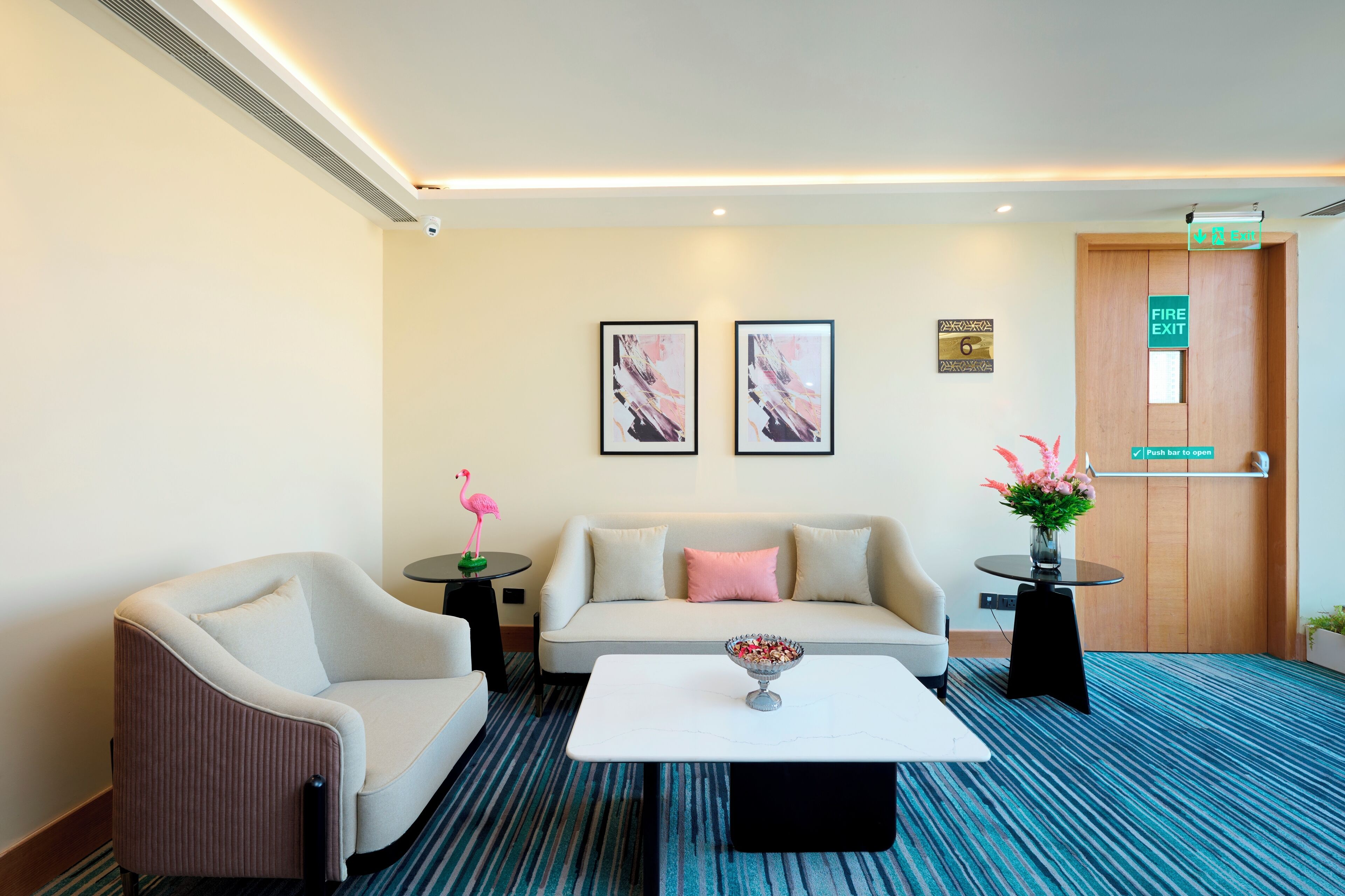 Een moderne hotelkamer met een bank, fauteuil en salontafel. Er hangen kunstwerken in lijsten aan de muur, een klein tafeltje met een bloemenvaas, en een bord met de tekst branddeur.
