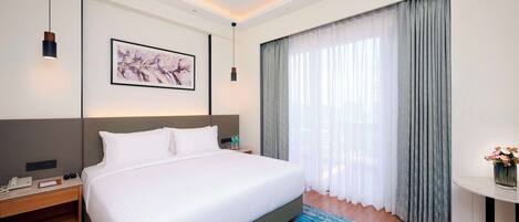 A hotel room with a large bed, a bedside table with a phone and a lamp, a framed picture on the wall, and a window with curtains.