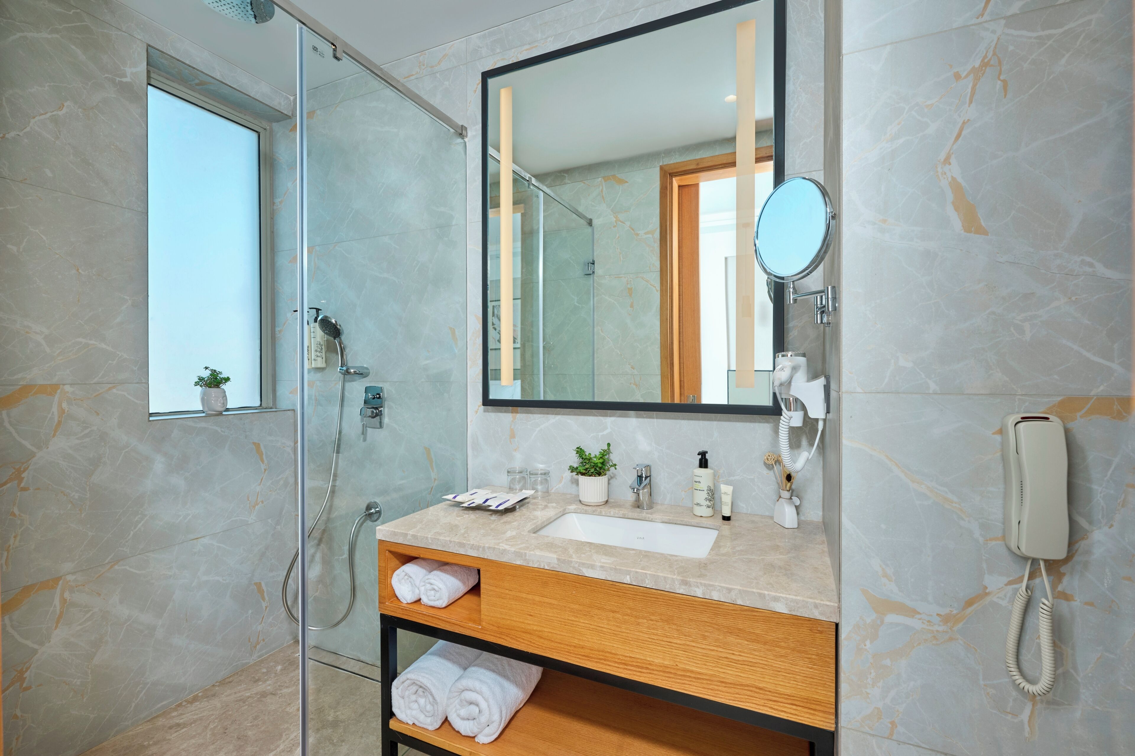 A modern bathroom with a glass-enclosed shower, a marble sink, a wooden vanity, and a mirror with a round frame.