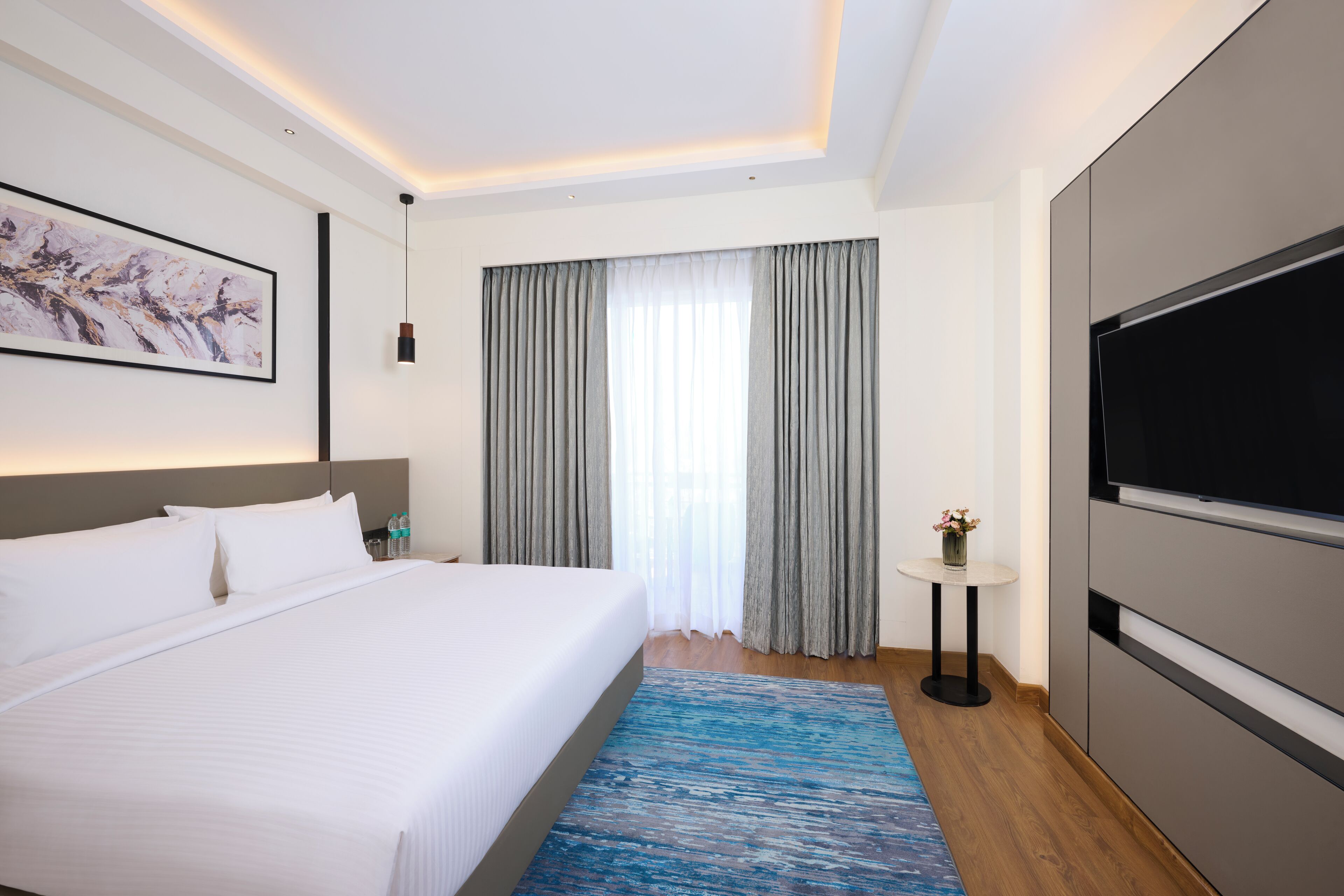A modern hotel room with a large bed, a flat-screen TV, a painting on the wall, and a window with curtains.