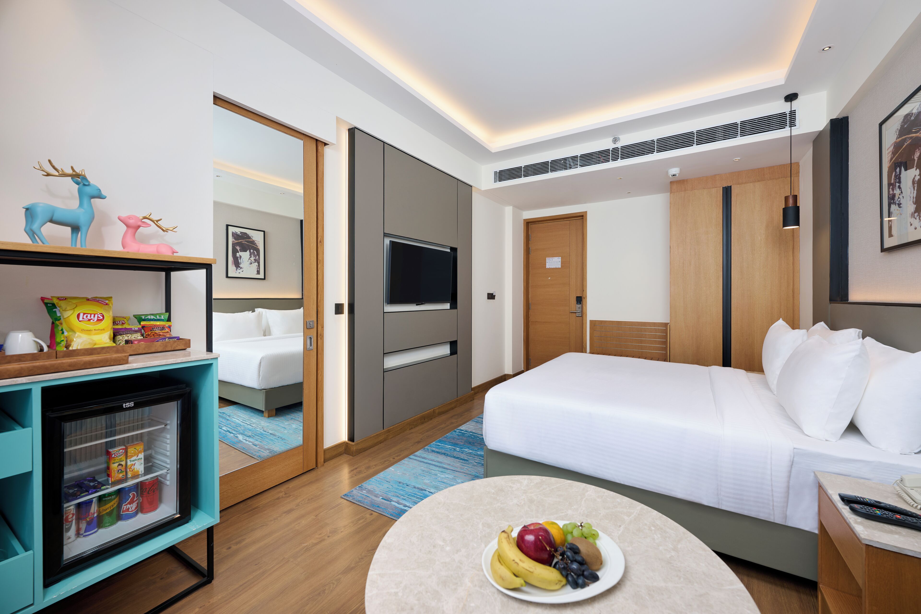 A modern hotel room with a bed, a television, a minibar, and a fruit bowl on the table.