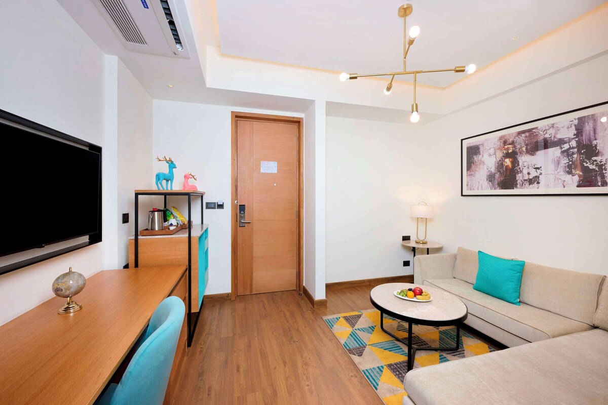 A modern hotel room with a wooden dining table, a sofa with a teal pillow, a round coffee table with fruit, a flat-screen TV, a wooden door, and a framed abstract painting on the wall.
