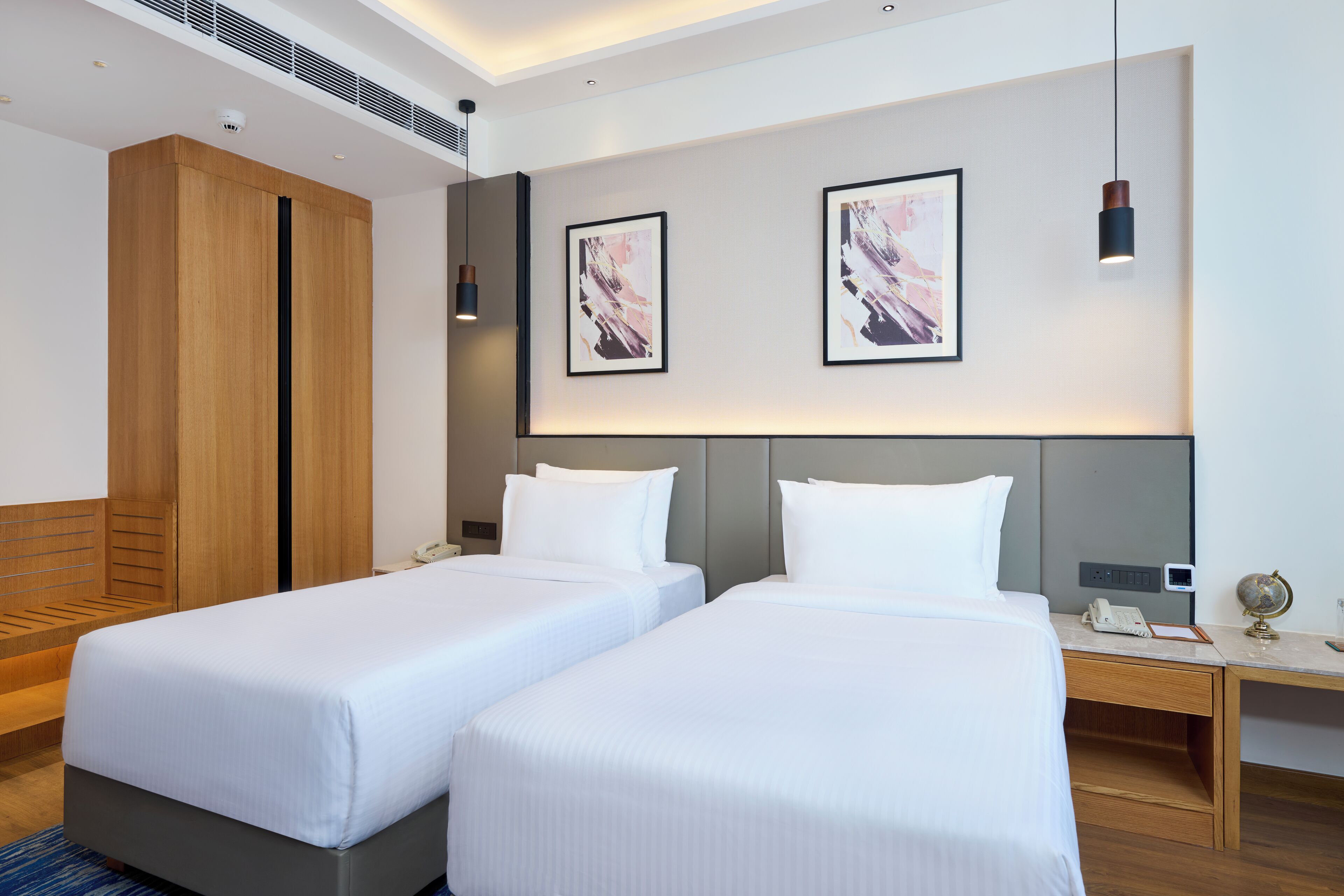 A hotel room with two single beds, a wooden wardrobe, a desk with a globe and phone, and abstract art on the wall.