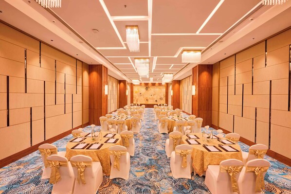 A long banquet hall with round tables, white chairs with gold sashes, and a patterned carpet. - Iris Sarovar Portico Sector 85 Gurugram (Gurugram)