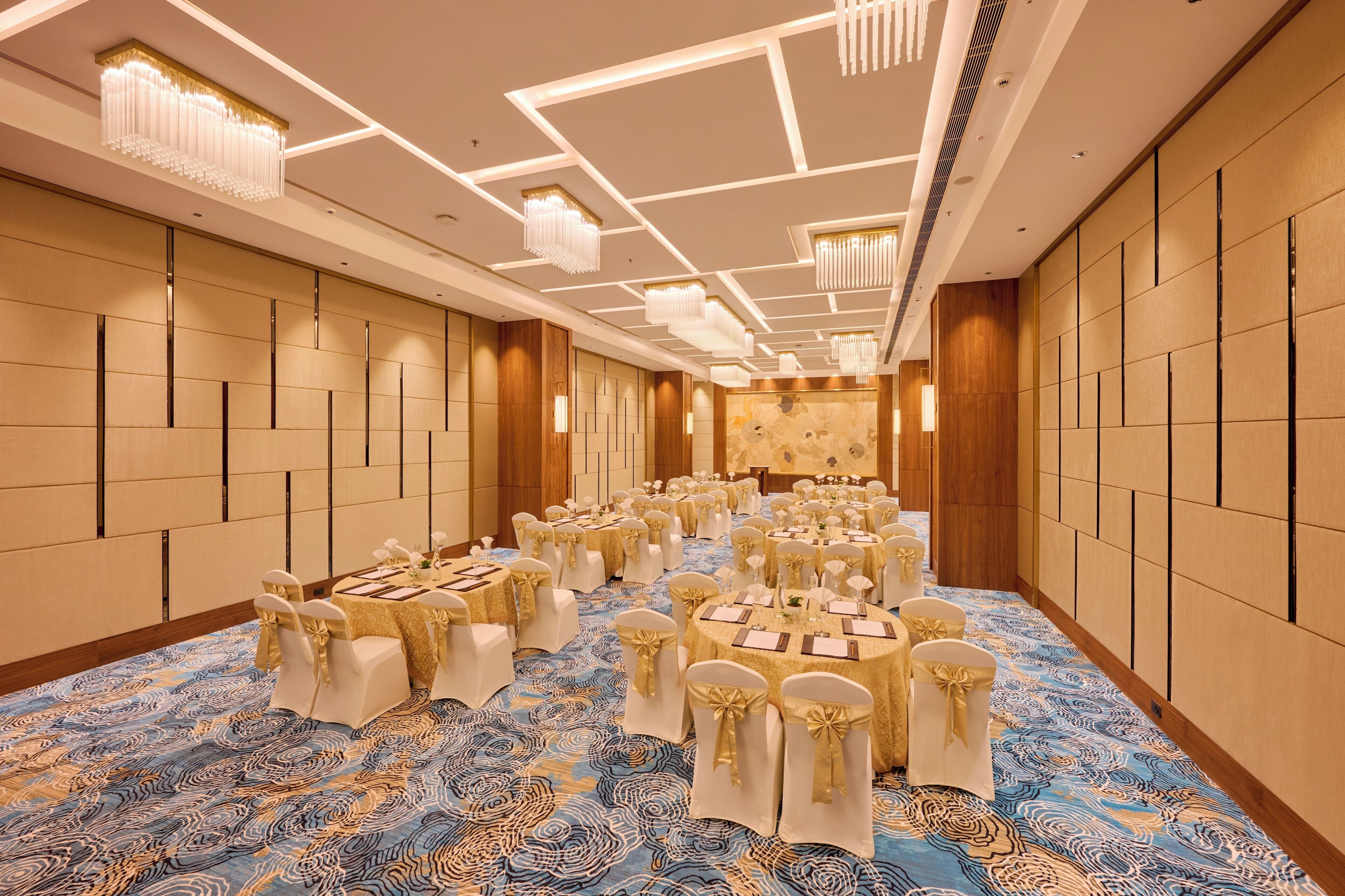 Une salle de banquet avec des tables rondes, des chaises blanches ornées de rubans dorés et un tapis à motifs.