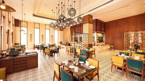 A spacious hotel dining area with multiple tables and chairs, a central bar area, and a large chandelier. - Iris Sarovar Portico Sector 85 Gurugram (Gurugram)