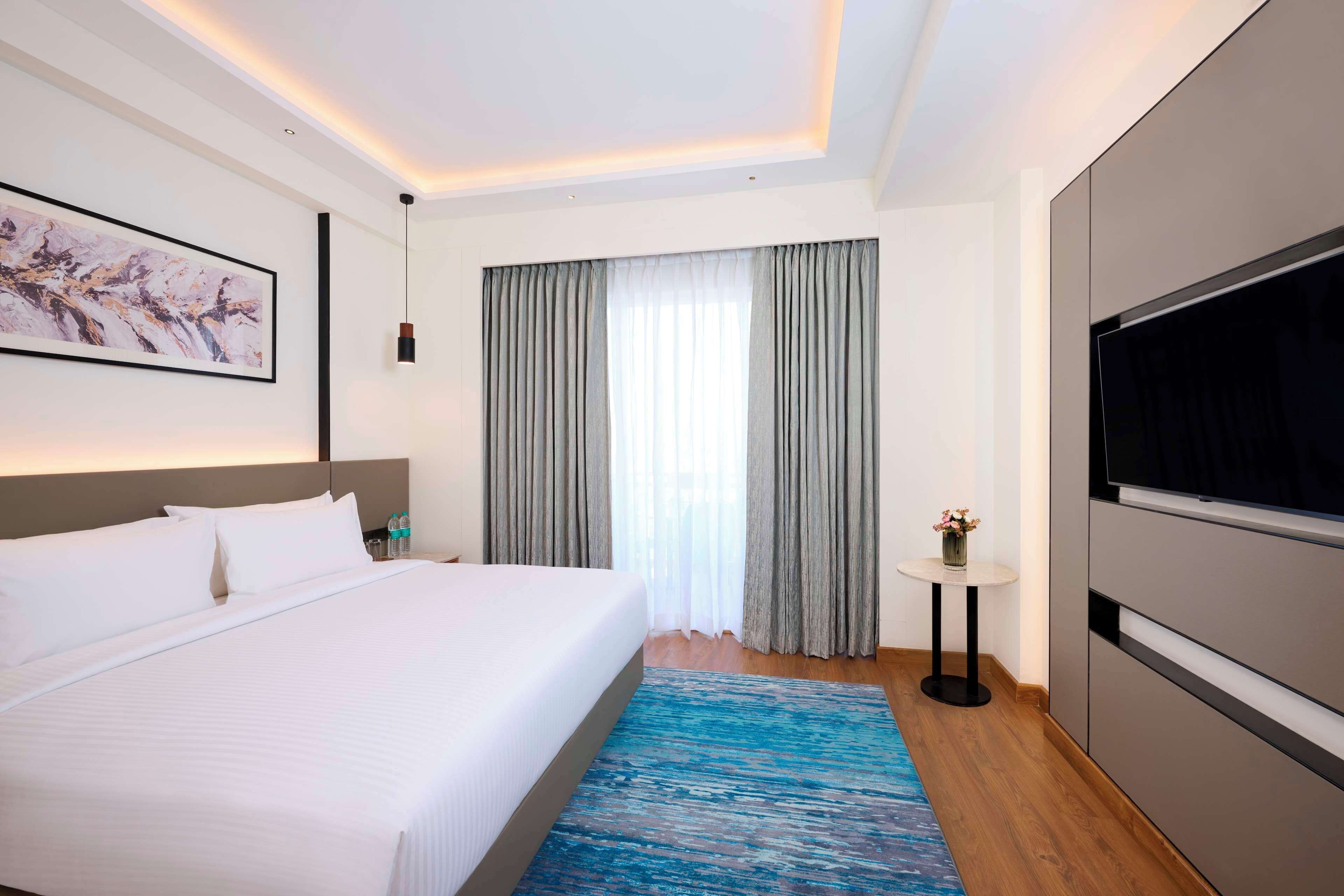 A modern hotel room with a large bed, a flat-screen TV, a painting on the wall, and a window with curtains.