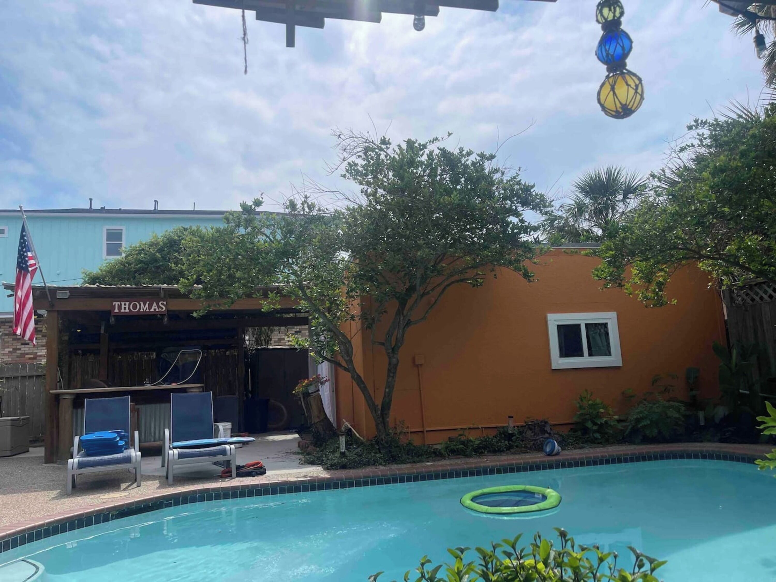 Una piscina con lettini, un piccolo edificio con la scritta 'THOMAS' e un albero sullo sfondo.