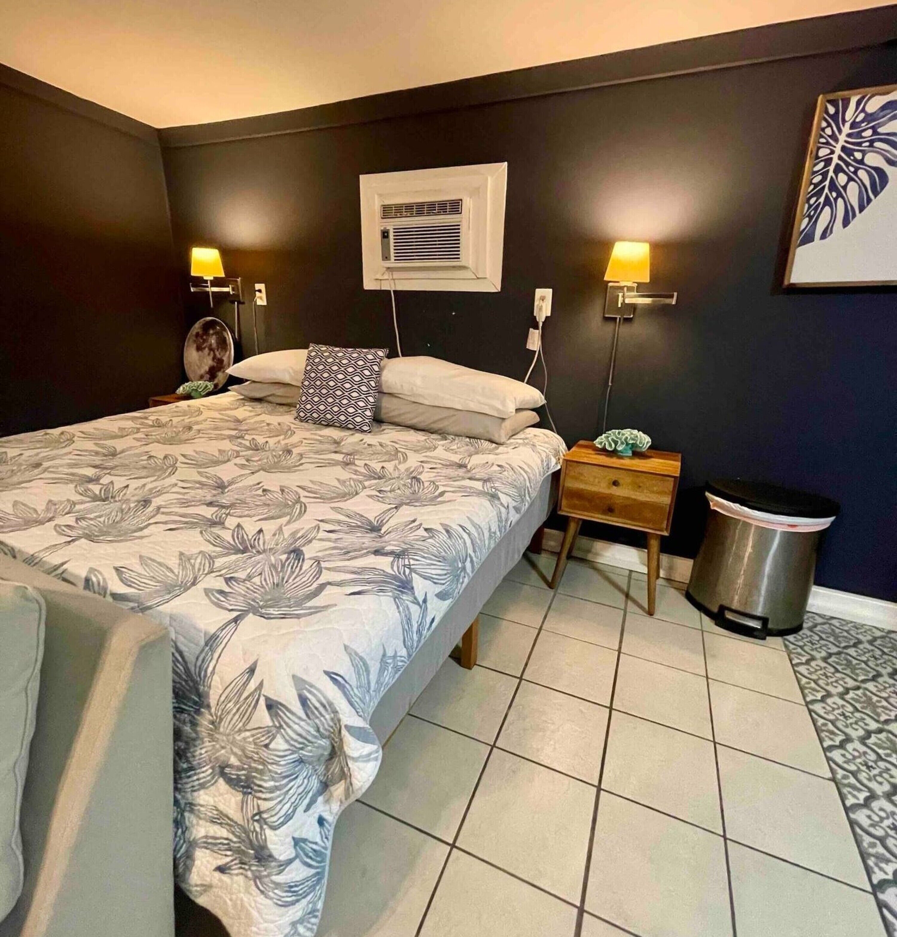 A bedroom with a bed, bedside lamps, a wall-mounted air conditioner, a trash bin, and a framed artwork on the wall.