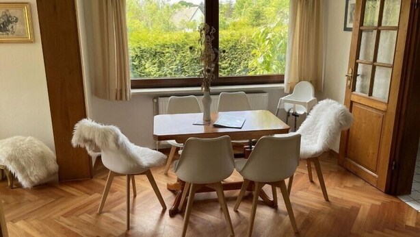 Un coin repas avec une table en bois, des chaises blanches, un lustre et une fenêtre avec des rideaux.