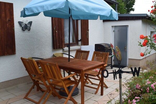 Un patio al aire libre con una mesa y sillas de madera, una sombrilla azul, una parrilla y una decoración con forma de mariposa.