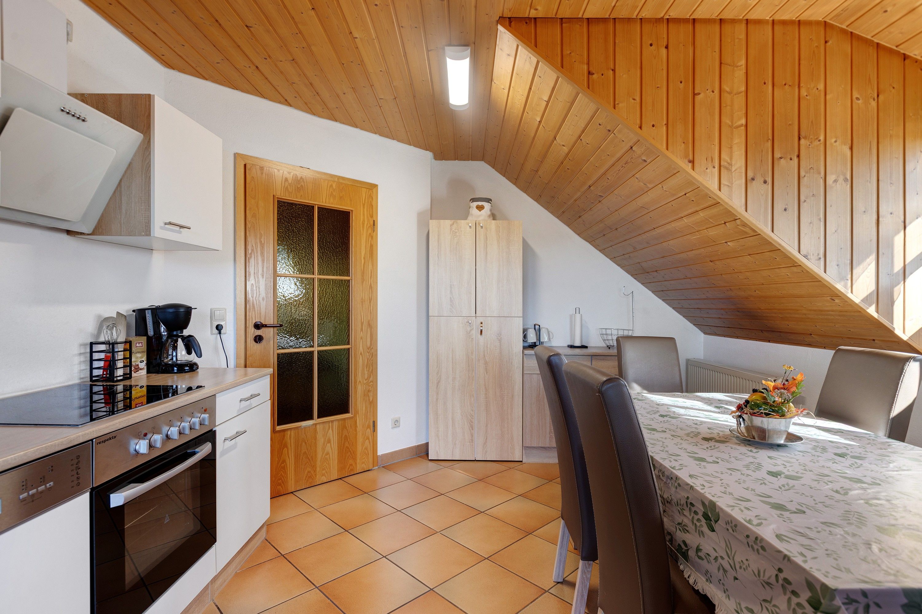 Een keuken met een fornuis, oven en kastjes, een eethoek met een tafel en stoelen, en een houten plafond.