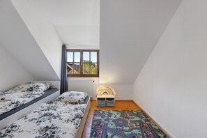 Une petite chambre mansardée, bien éclairée, avec deux lits, une fenêtre et un tapis coloré.