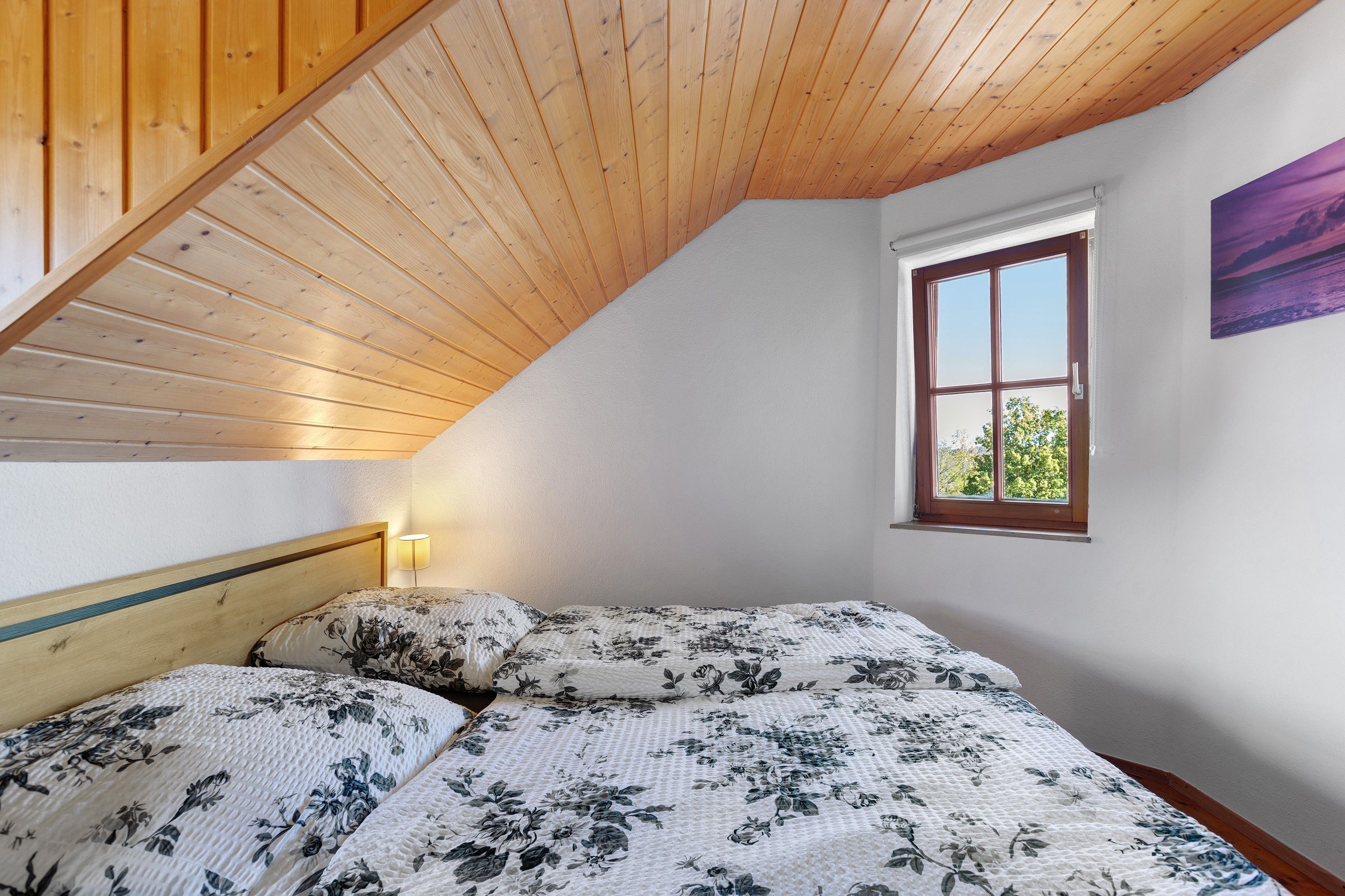 Ein Schlafzimmer mit schrägverdeckter Decke, einem Bett mit floralem Bettzeug, einer Nachttischlampe und einem Fenster mit Aussicht.