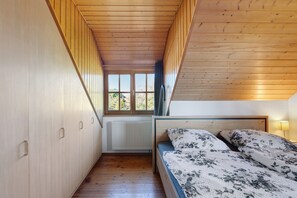 Een kleine kamer met een schuine houten balkenplafond, een bed met bloemenbedrukking en een raam met uitzicht op groen.