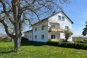 Ein zweigeschossiges weißes Gebäude mit Balkon, umgeben von einer Rasenfläche und einem großen Baum.