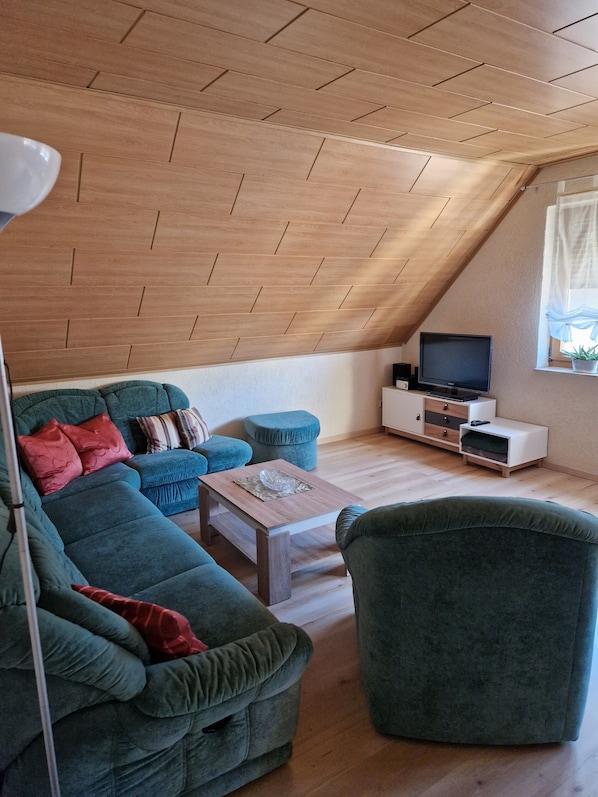 Ein Loft-Zimmer mit Holzbalkendecke, einer Sofagarnitur, einem Couchtisch, einem Fernseher und einem Fenster mit Vorhängen.