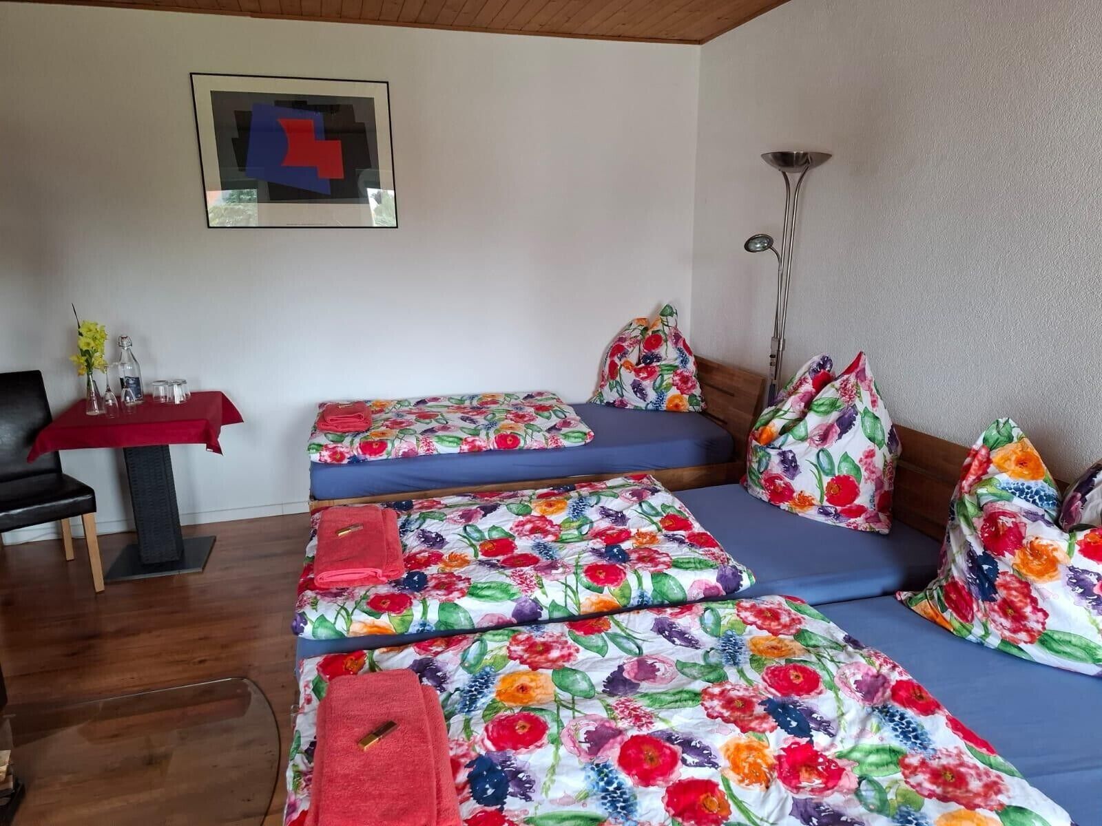 Quarto com duas camas, uma mesa com toalha vermelha, uma cadeira e um abajur.