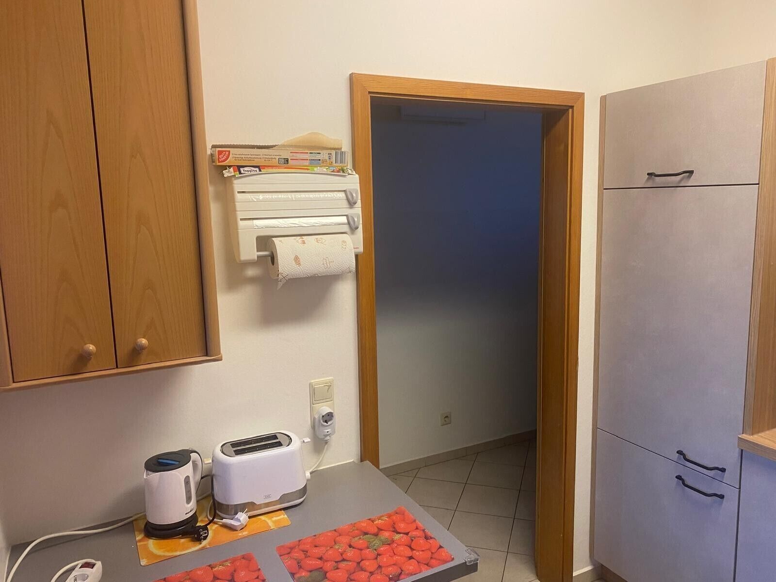 Une petite cuisine équipée d’un grille-pain, d’une bouilloire et d’un plateau de tomates. On y trouve des placards en bois et une porte qui donne sur une autre pièce.