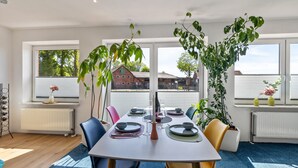 Uma sala de jantar com mesa posta para quatro pessoas, rodeada por cadeiras, janelas amplas e plantas em vasos.