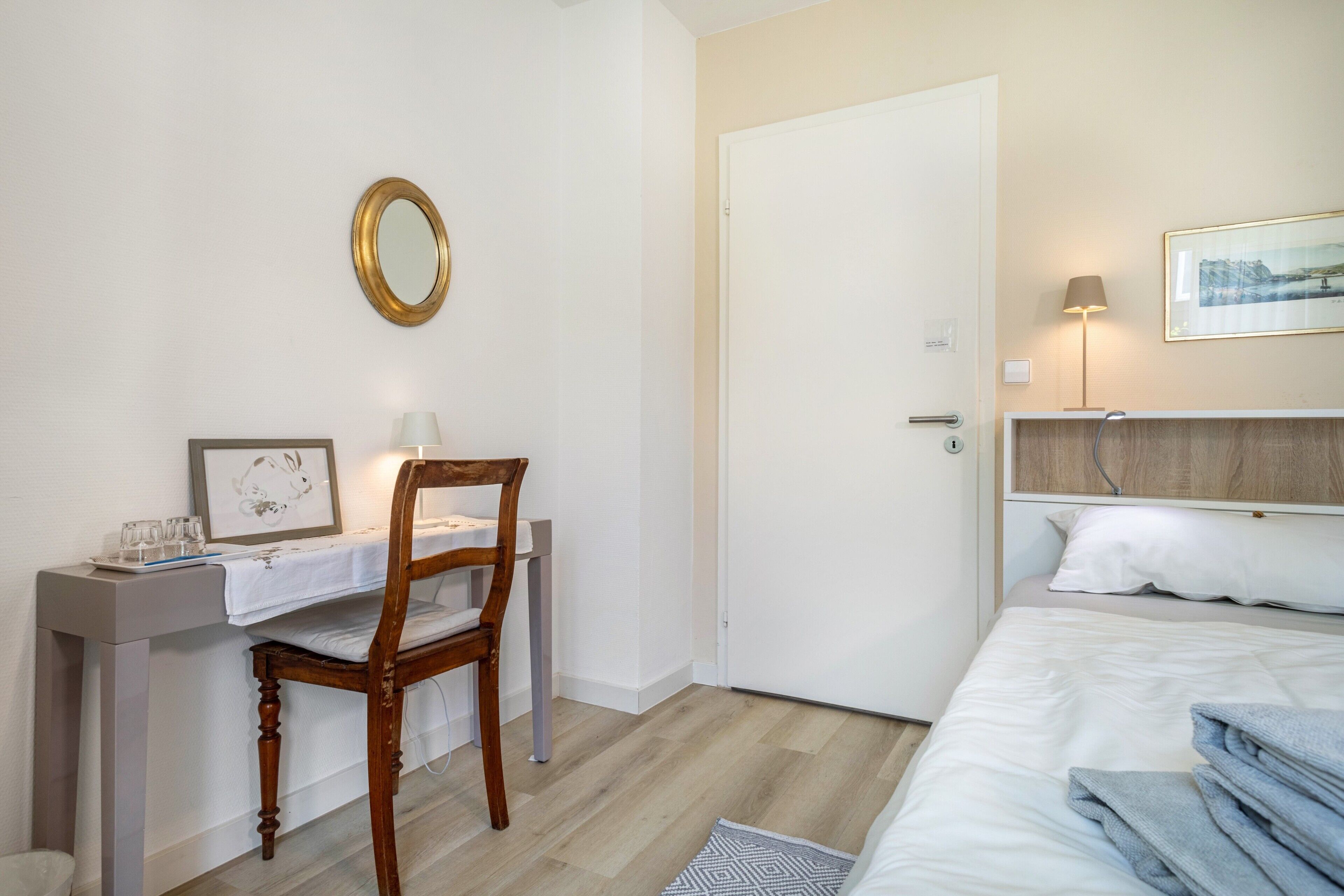 Ein Schlafzimmer mit einem Bett, einem Schreibtisch mit Stuhl, einem Nachttisch, einer Tür, einem an der Wand befestigten Spiegel und einem gerahmten Bild.
