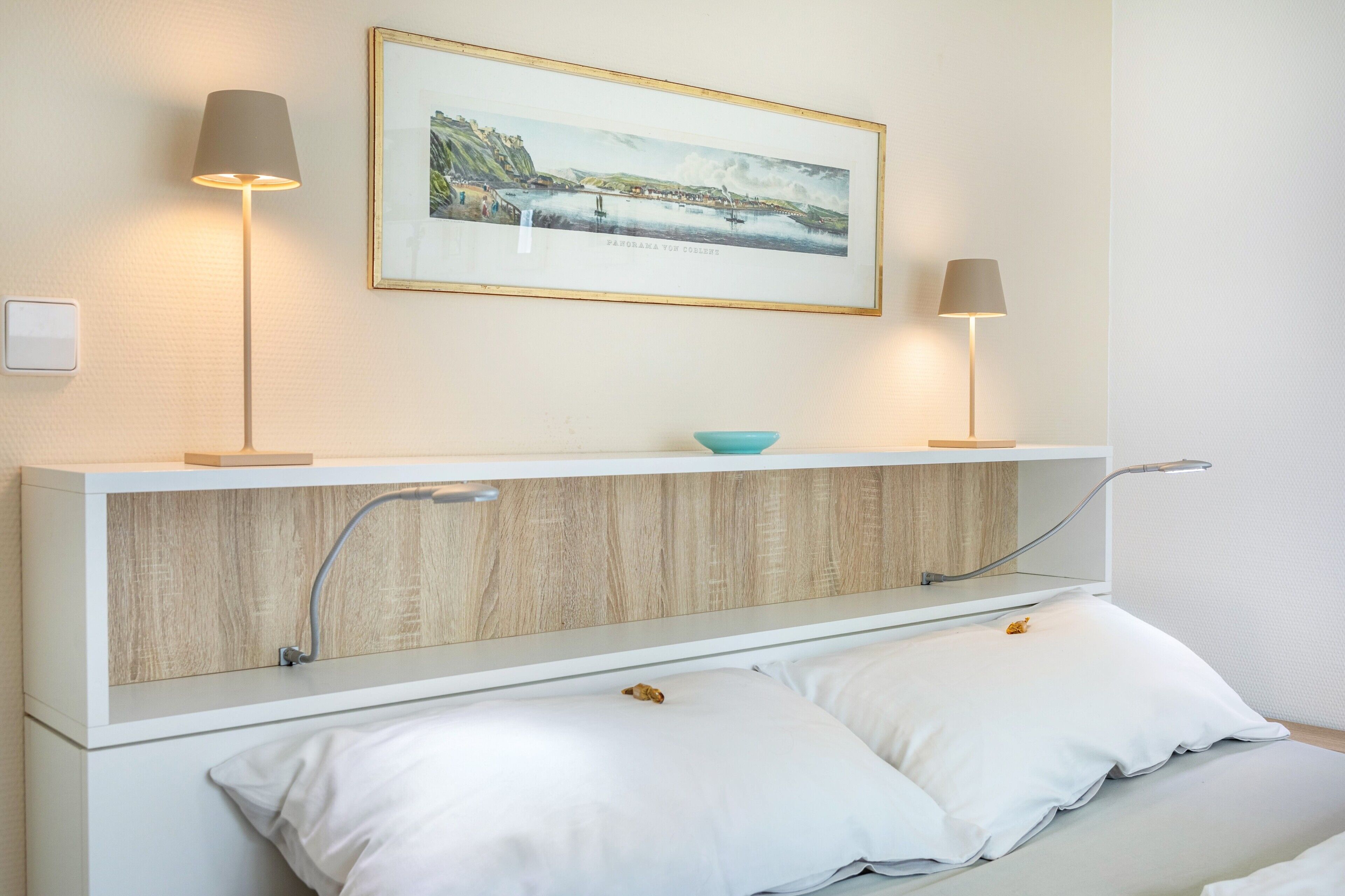 Une chambre à coucher moderne avec un lit, deux lampes de chevet, un tableau encadré et une petite étagère sur laquelle se trouve un bol.