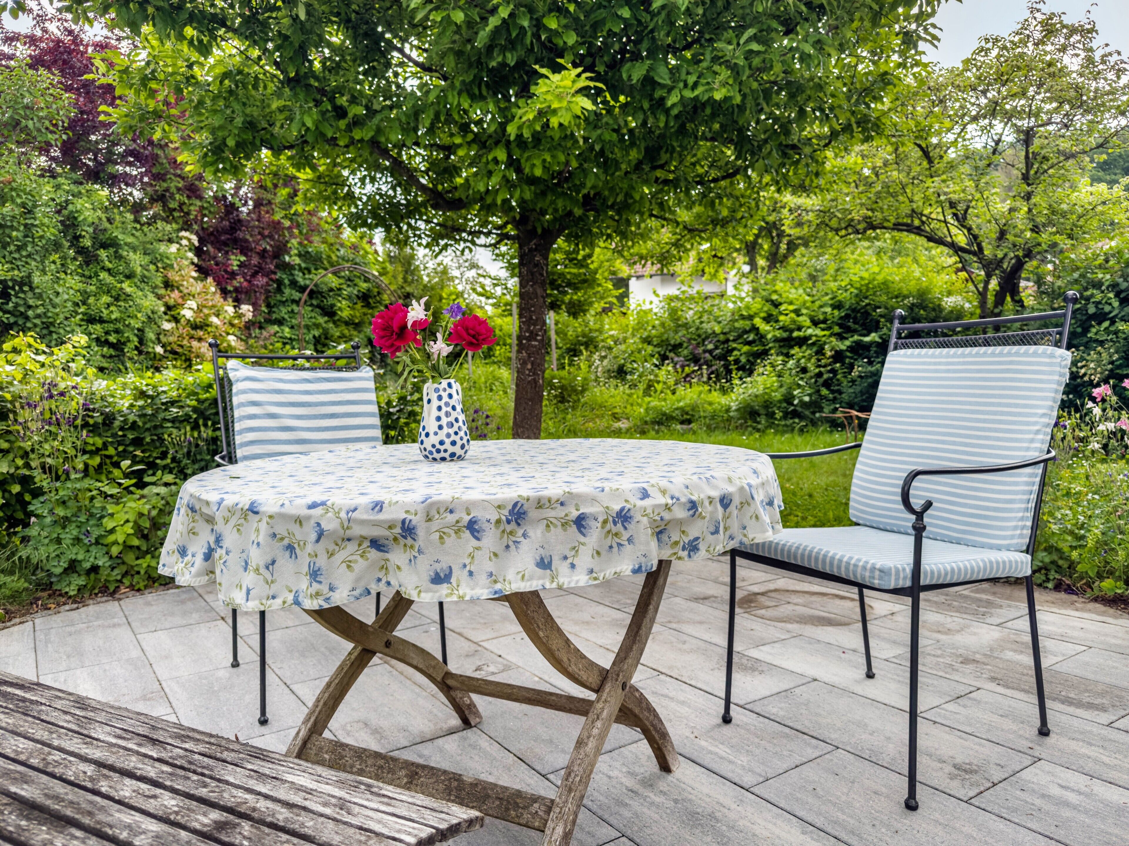 Een ronde tafel met een tafelkleed met bloemen, twee stoelen met gestreepte kussens en een vaas met bloemen.