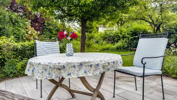 Een ronde tafel met een tafelkleed met bloemen, twee stoelen met gestreepte kussens en een vaas met bloemen.