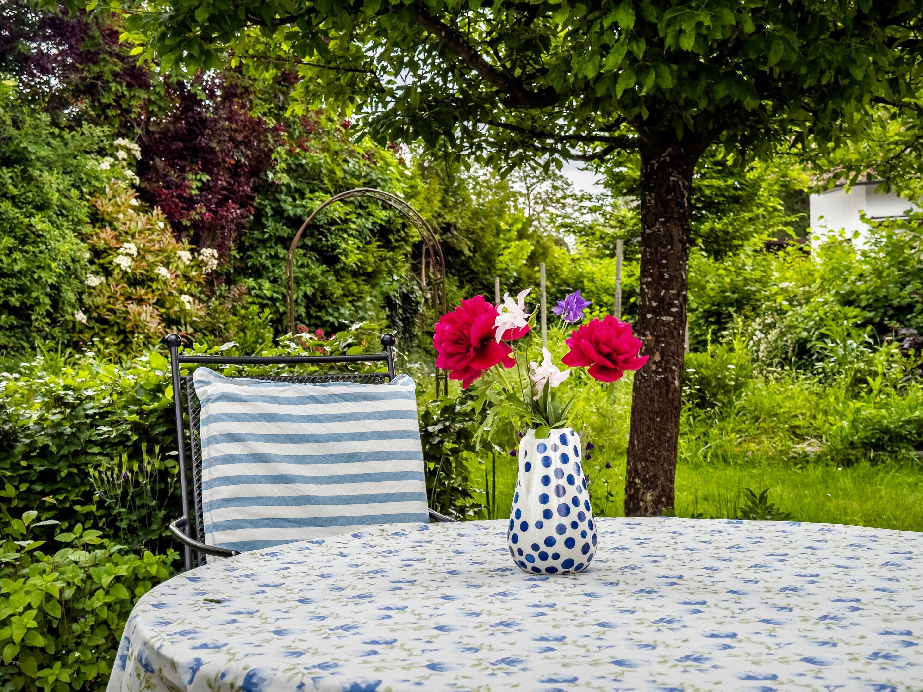 A round table with a floral tablecloth, a polka-dotted vase with flowers, and a striped cushion on a metal chair.