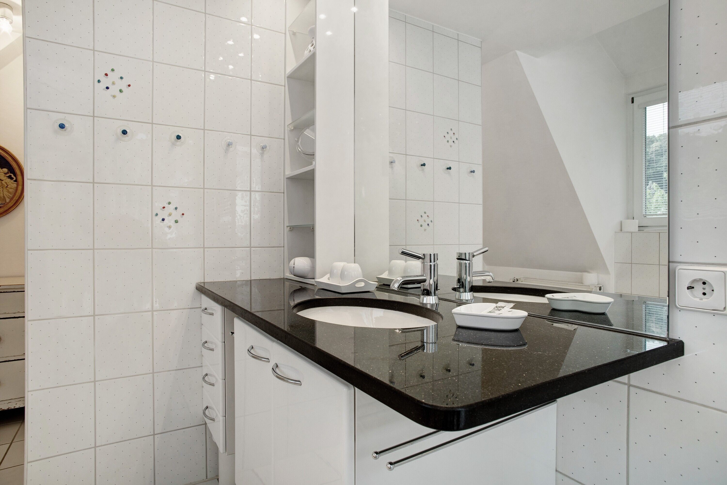 Un baño moderno con paredes de azulejos blancos, encimera negra, dos lavamanos, un espejo grande y una ventana.