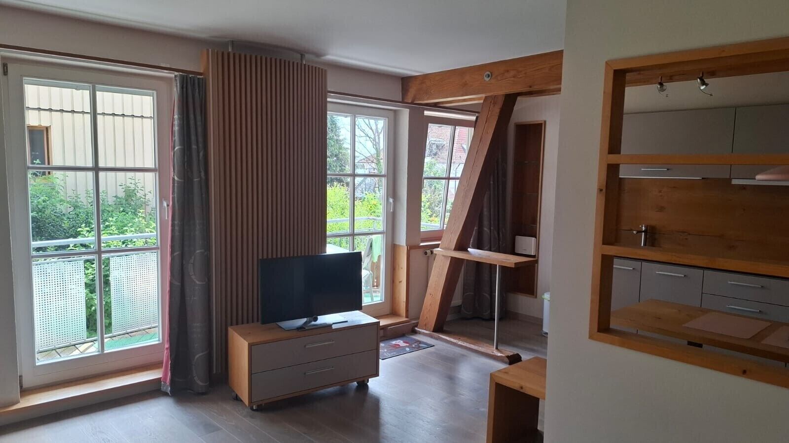 A room with a wooden TV stand, a large window with curtains, and a wooden beam structure.
