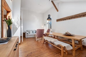 A wooden dining table with benches, a sofa, and a chair in a bright room with wooden floors and white walls.