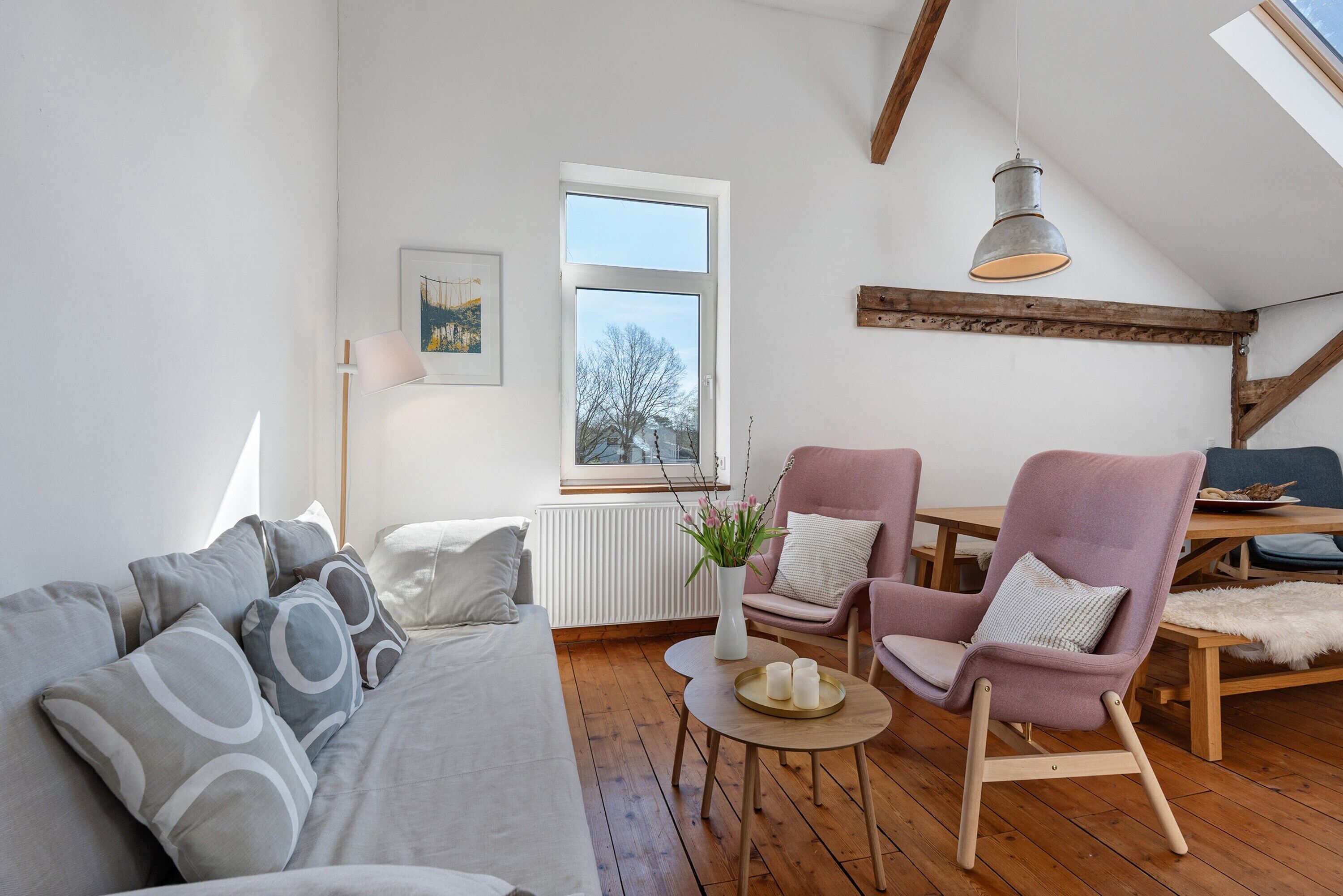 A modern loft-style room with a wooden floor, a sofa, two armchairs, a small round table with a vase and candles, and a large window with a view of trees.
