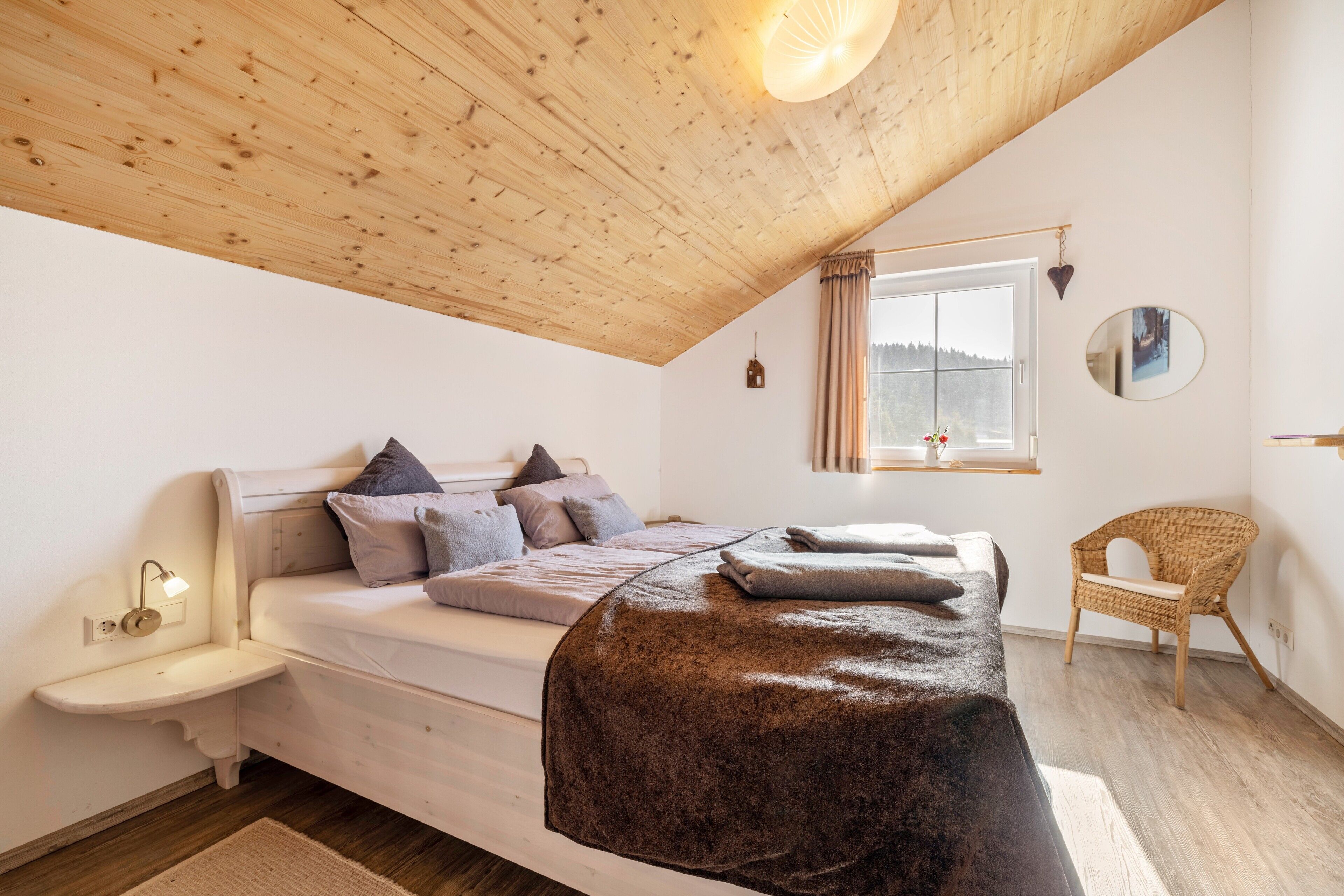 Un dormitorio con techo de madera, una cama con almohadas, una mesita de noche, una silla y una ventana con cortinas.