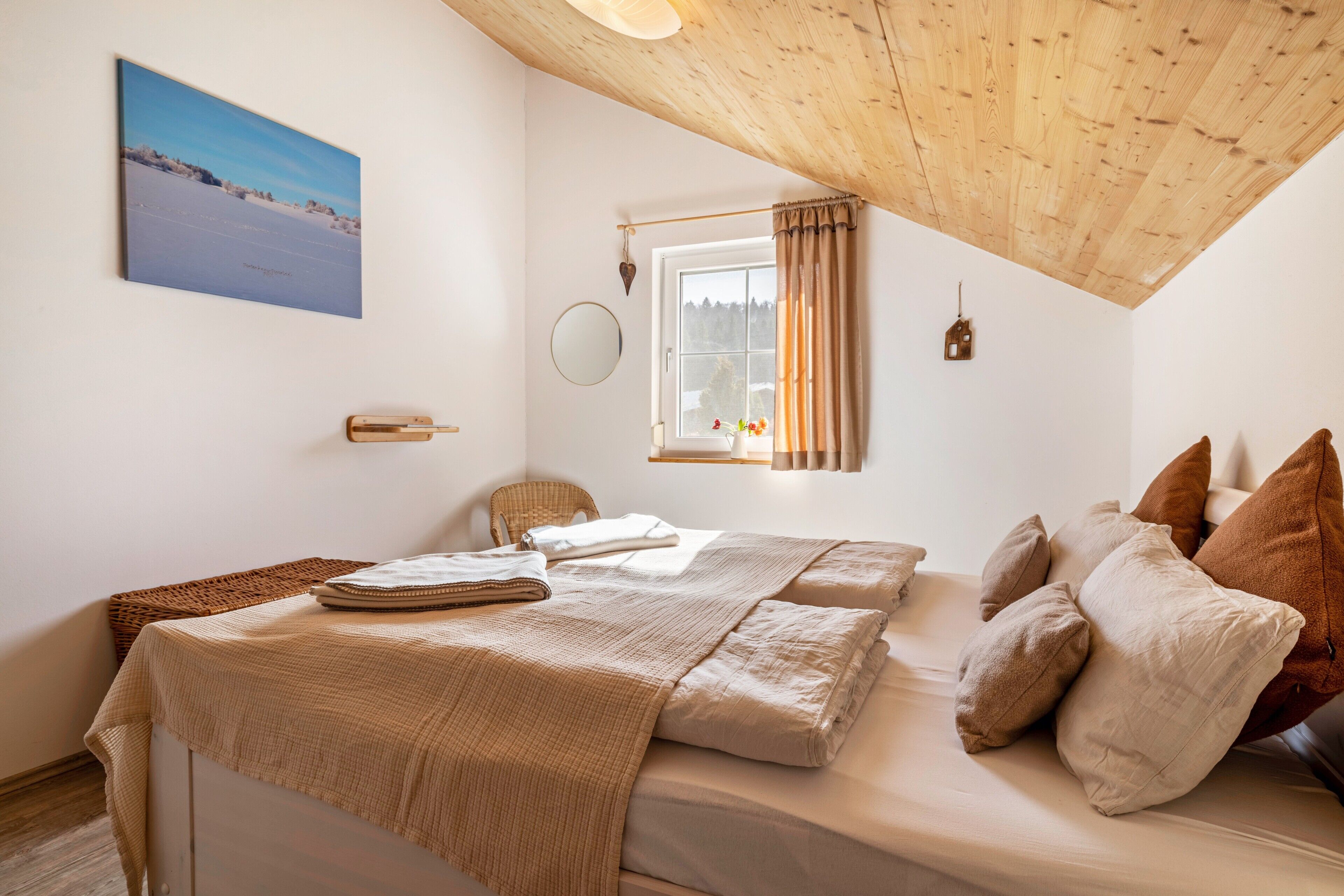 A bedroom with a wooden ceiling, a bed with beige bedding, a window with curtains, a wall-mounted mirror, and a framed picture.
