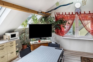Habitación con televisión, microondas, mesa de comedor, sillas, plantas en macetas y ventana con cortinas.