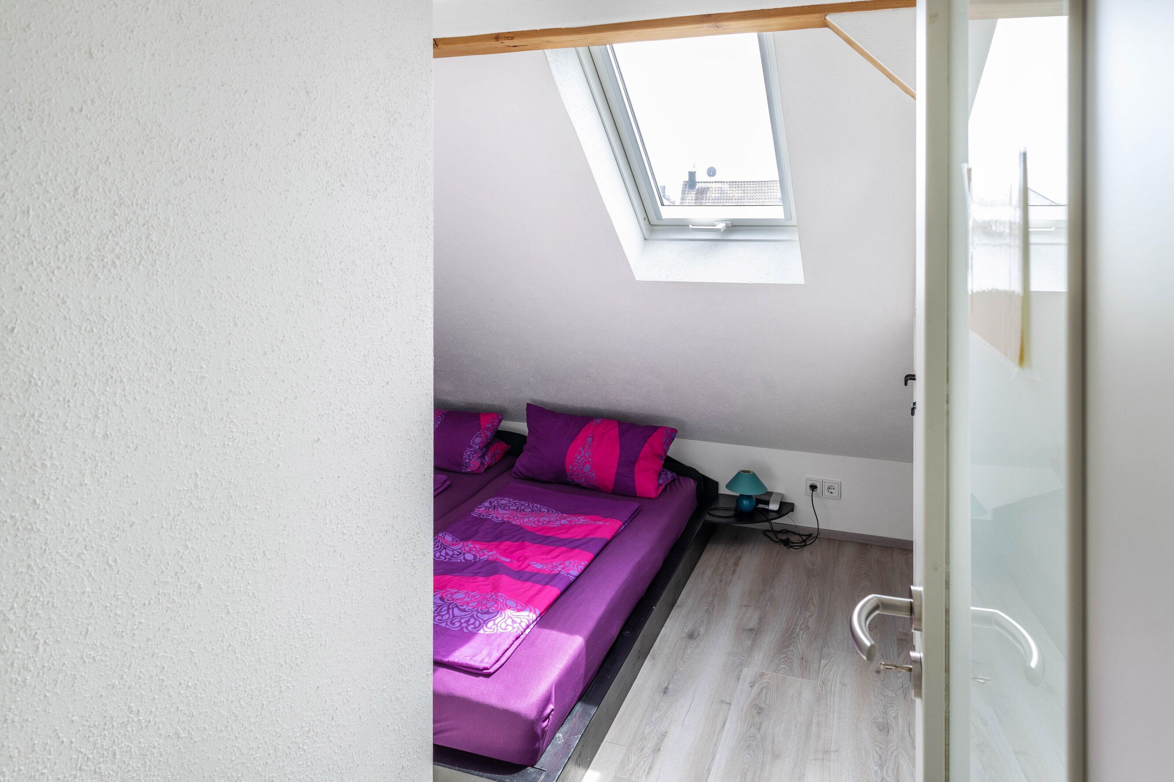 A bedroom with a skylight, a bed with purple bedding, a nightstand with a lamp, and a wooden floor.