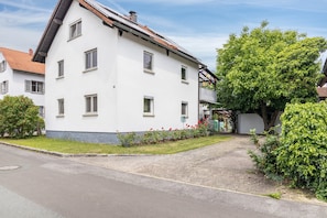 En toetages hvid bygning med skråt tag, en belagt indkørsel og en velplejet have med røde blomster.