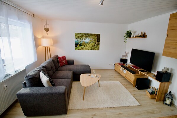 A living room with a sectional sofa, a small round coffee table, a flat-screen TV, a wooden sideboard, and a framed landscape painting.