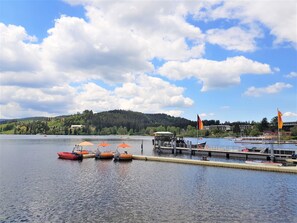 Ein See mit Booten an einem Steg, einer Flagge und einem Hügel mit Bäumen und Gebäuden im Hintergrund.